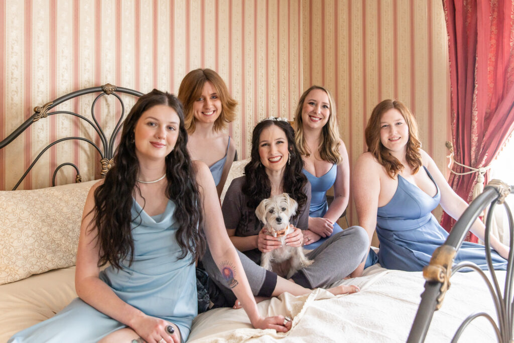 A bride sits on a bed surrounded by members of her wedding party while holding a small white dog in her lap. The dog looks toward the camera as bridesmaids in light blue dresses gather around, creating a relaxed getting-ready portrait that includes an important member of the family.