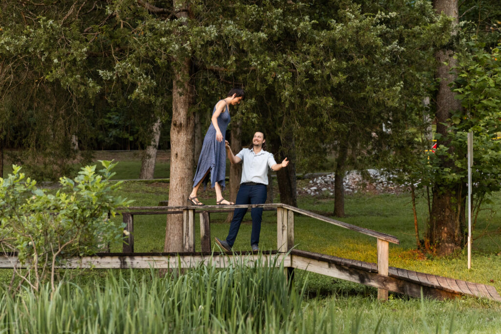 During an engagement session, one partner carefully balances on a wooden bridge while the other reaches up with open arms, surrounded by tall trees and lush greenery. Working with LGBTQ friendly wedding vendors often means feeling free to express your personality and relationship naturally