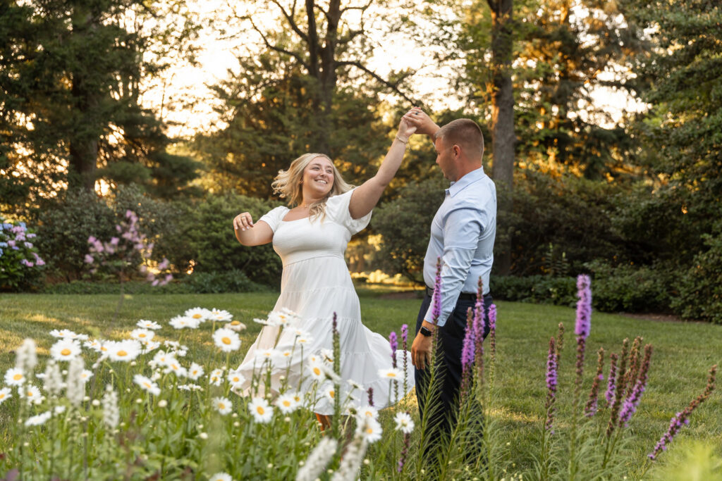 A couple dances among blooming daisies and purple flowers during golden hour, with warm sunlight filtering through the trees during their Drumore Estates engagement session.
