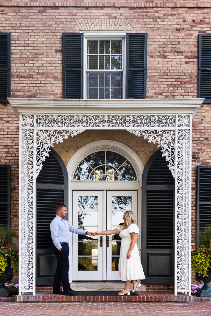 The couple stands beneath the ornate white ironwork entrance of the estate during their engagement, reaching for each other's hands in front of the historic brick façade that makes a Drumore Estates wedding so distinctive.