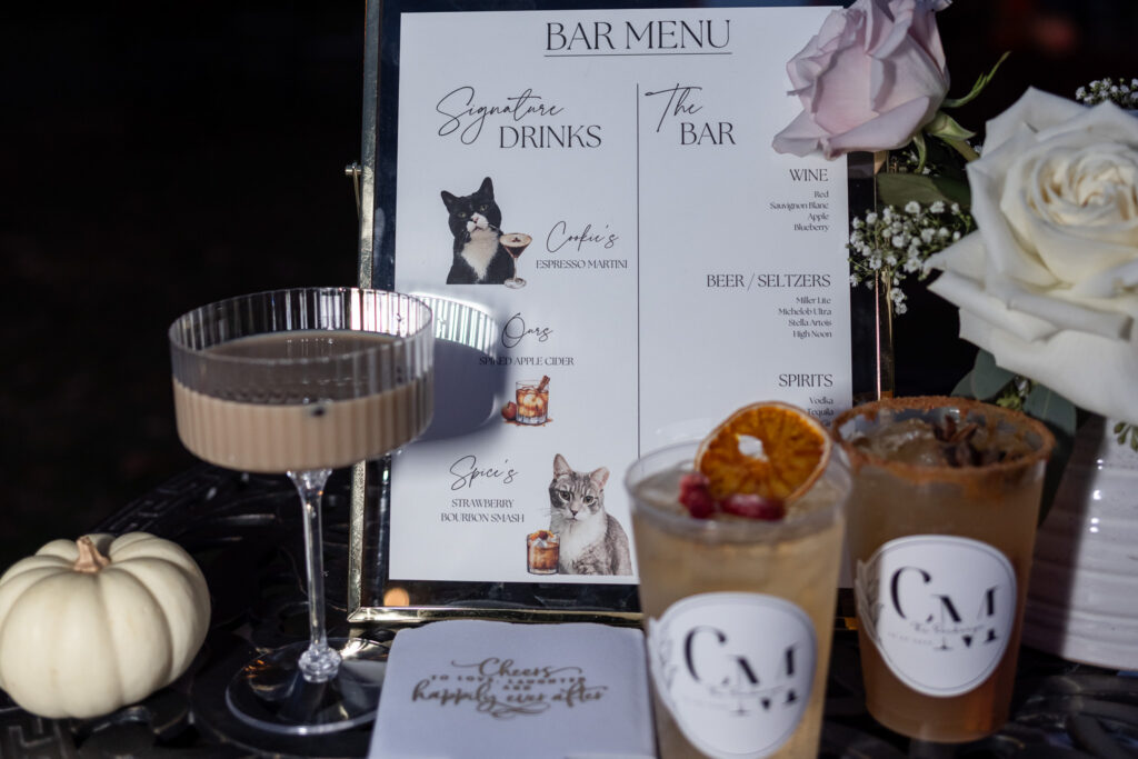 Wedding reception detail photograph featuring signature cocktails named after the couple's cats. Custom illustrations of the pets appear on a framed bar menu surrounded by drinks, florals, and seasonal décor, offering a creative way to include pets who could not attend the celebration.