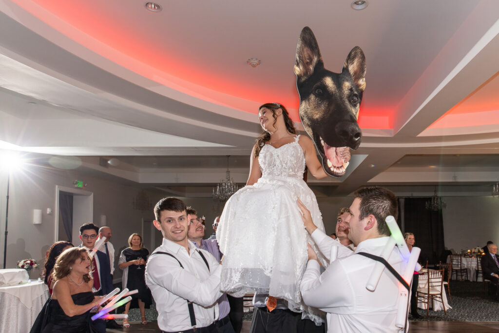 A bride is lifted high above the dance floor during the reception while holding a giant cutout of her dog's head. Guests cheer and wave glow sticks around her, turning the oversized pet portrait into a fun and memorable reception moment.
