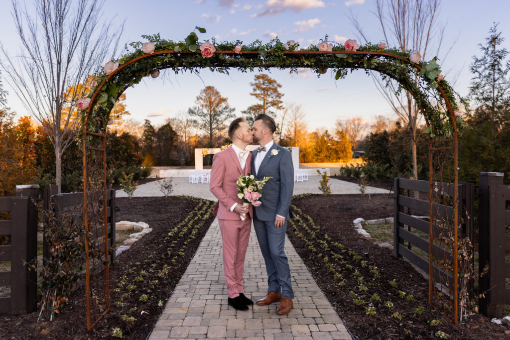 Two grooms stand beneath a flower-covered arbor at sunset, one dressed in pink and holding a bouquet. They share a kiss while surrounded by landscaped gardens and soft golden light. Finding LGBT friendly wedding vendors can help ensure your wedding day feels supportive from beginning to end