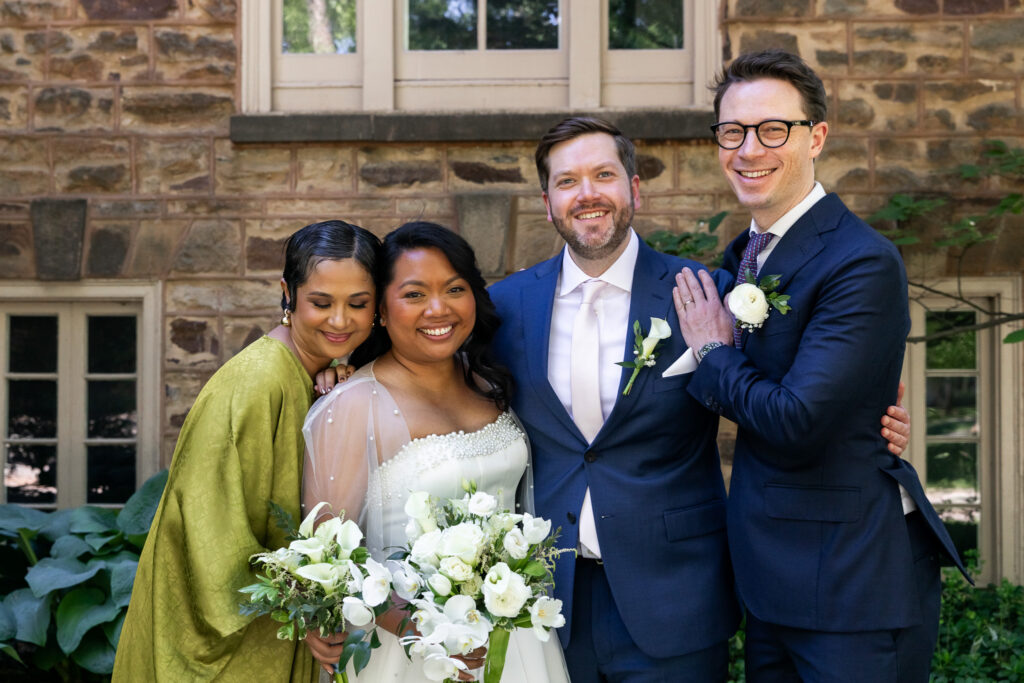 Bride, groom and their wedding party come in for a hug in front of a historic stone building 
