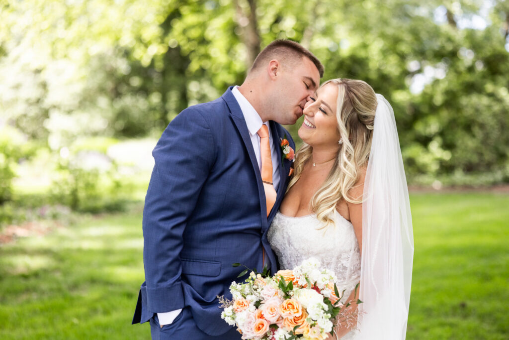 A groom in a blue suit leans in to kiss his bride's cheek as she smiles and holds a bouquet of peach and ivory roses, creating a joyful portrait from their Drumore Estates wedding surrounded by lush garden greenery and soft natural light.