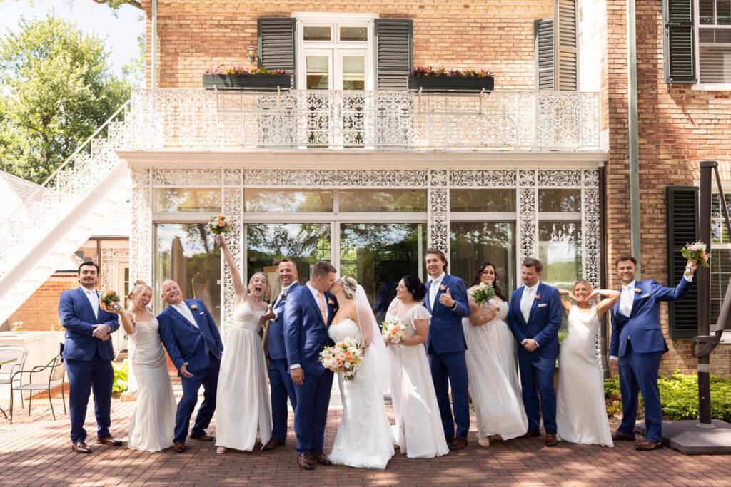 Playful wedding party portrait in front of an elegant brick estate, with the newlyweds sharing a kiss at the center while bridesmaids, groomsmen, and family members strike fun poses and celebrate around them.