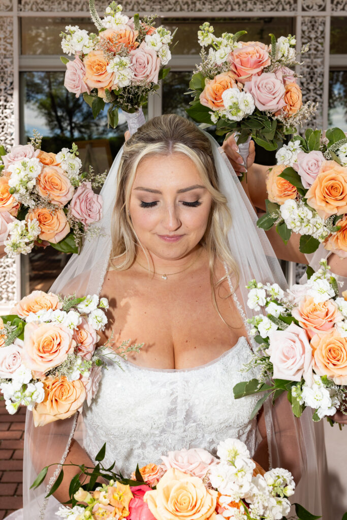 Bridal portrait featuring the bride surrounded by bouquets of peach, blush, and white flowers held by her wedding party. Soft florals frame her face and veil as she looks down with a calm expression.