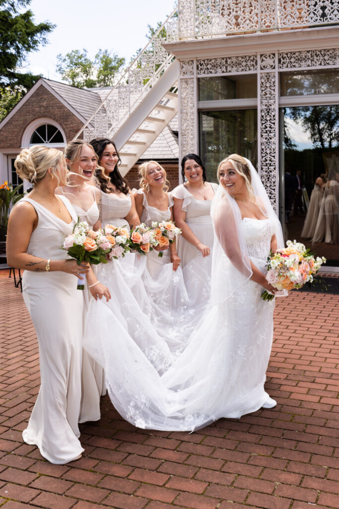 Bride and bridesmaids laugh together during wedding party portraits outside the venue. Flowing dresses, pastel bouquets, and the bride’s long veil create a joyful and lighthearted scene.