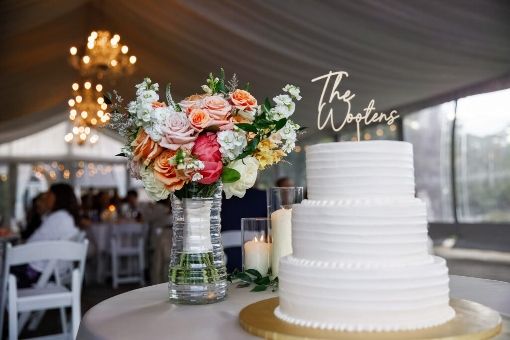 A three-tier white wedding cake sits beside a colorful arrangement of peach, blush, coral, and white flowers during a Lancaster Wedding reception. A custom acrylic topper displaying the couple’s last name rises above the cake beneath elegant tent lighting.