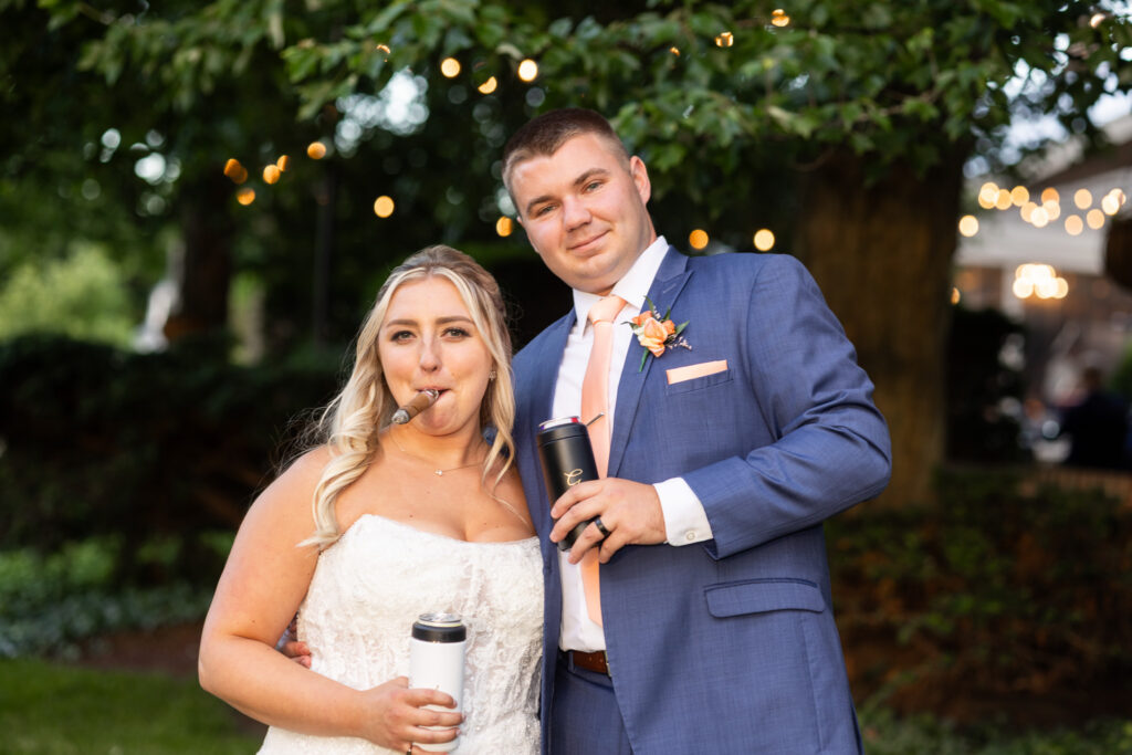 Newlyweds pose together during their Lancaster Wedding reception, holding insulated drink tumblers while the bride playfully pretends to smoke a cigar. Warm string lights and greenery create a relaxed evening atmosphere.