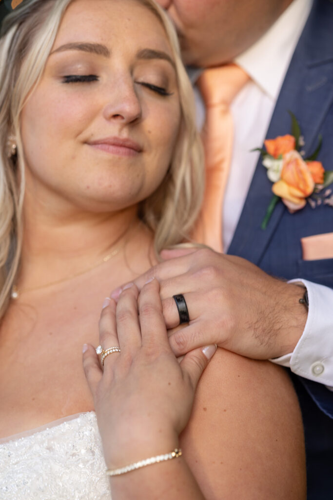 Close-up wedding portrait showing the bride’s engagement ring and wedding band resting on her shoulder while the groom embraces her from behind. Soft natural light highlights the details of their rings and attire.
