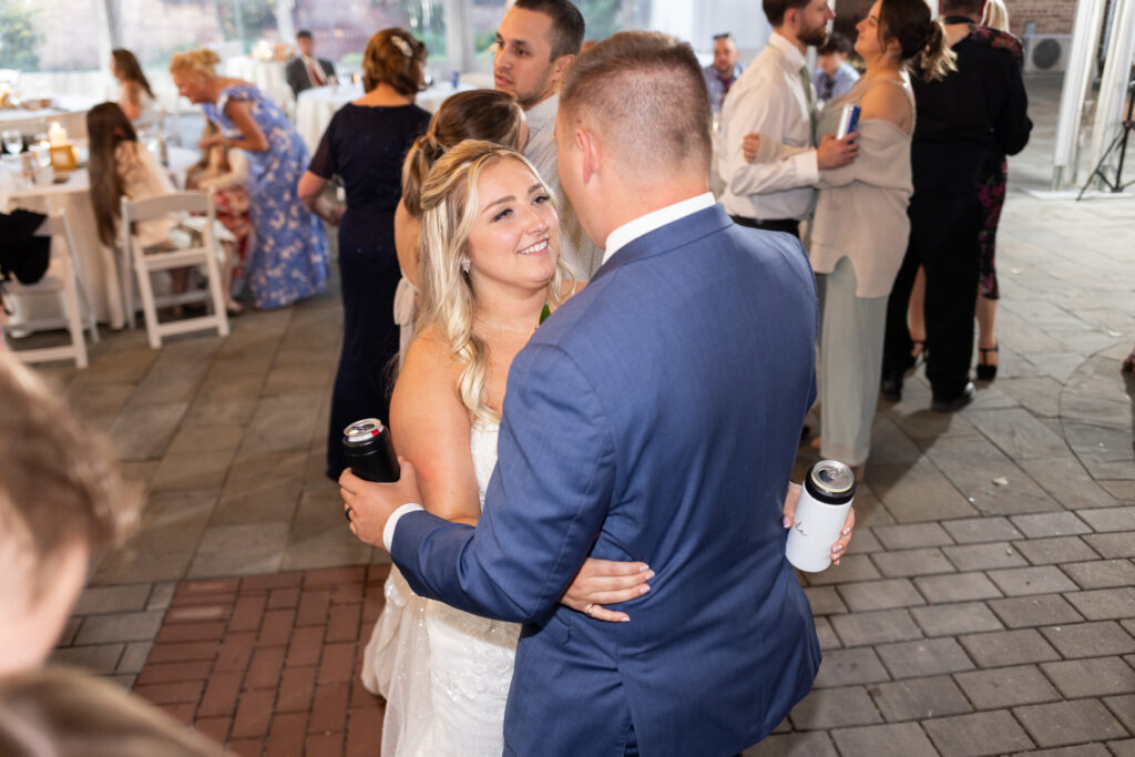Bride and groom share a close dance during their Lancaster Wedding reception, holding personalized drink tumblers while smiling at one another. Guests and family members gather nearby as the celebration continues around them.