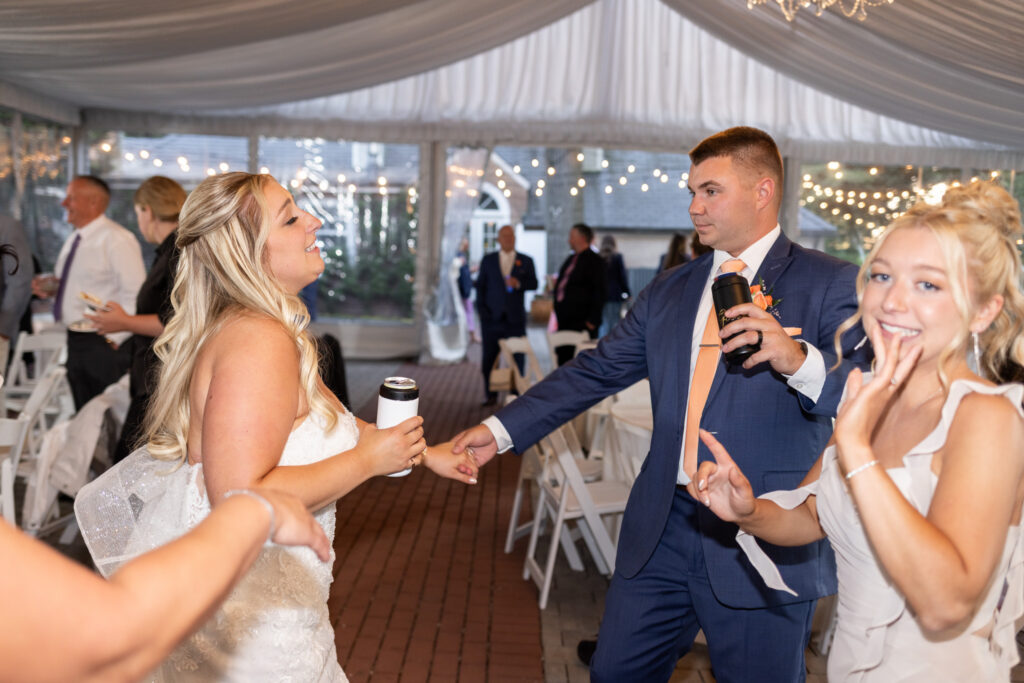 The bride and groom dance and laugh with friends and family during a lively Lancaster Wedding reception under a tent filled with twinkling lights and white drapery.