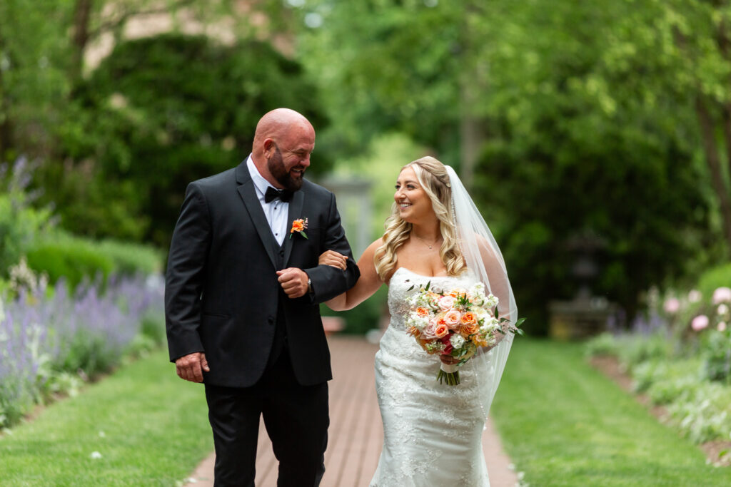 A bride walks arm-in-arm with her father down a garden pathway, carrying a peach and ivory bouquet as they make their way toward the ceremony.