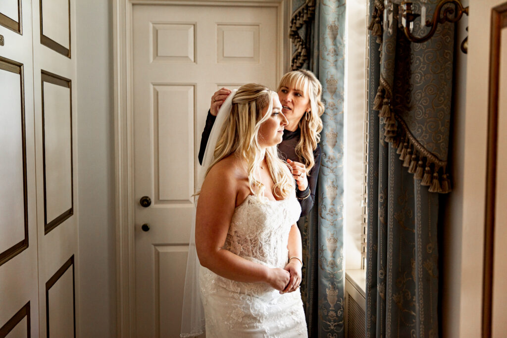 A loved one adjusts the bride’s veil near a window as natural light falls across her wedding gown. The quiet getting-ready moment highlights the final touches before she walks down the aisle.