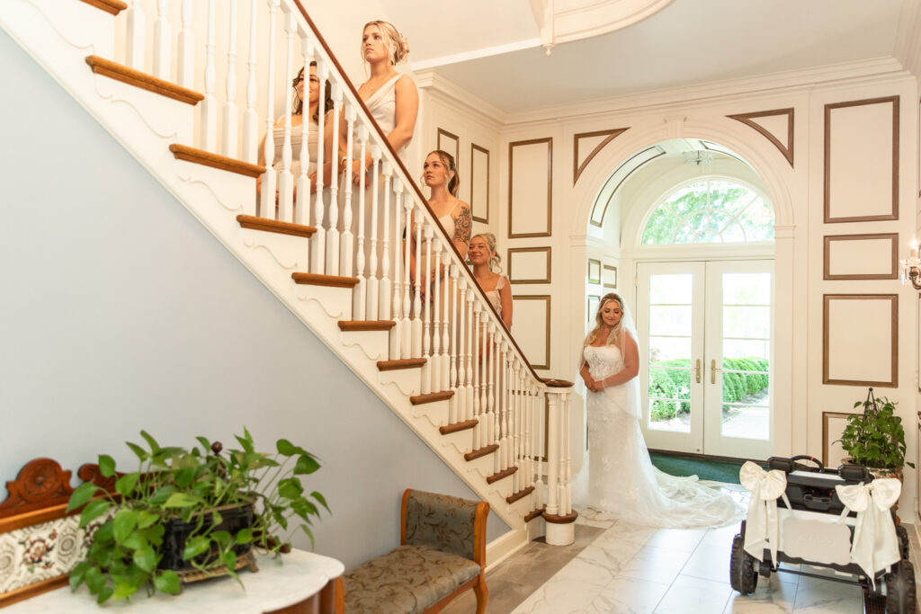 Bridesmaids line a grand staircase while the bride waits below in the foyer of the wedding venue for their first look. The scene showcases coordinated dresses, elegant architecture, and anticipation before the ceremony.
