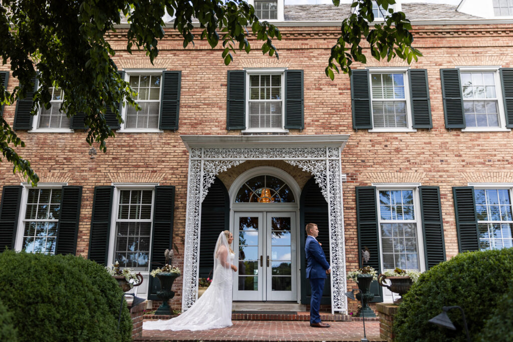 The bride approaches her groom from behind for their first look outside the historic manor, framed by manicured hedges and the iconic entrance of a Drumore Estates wedding venue.