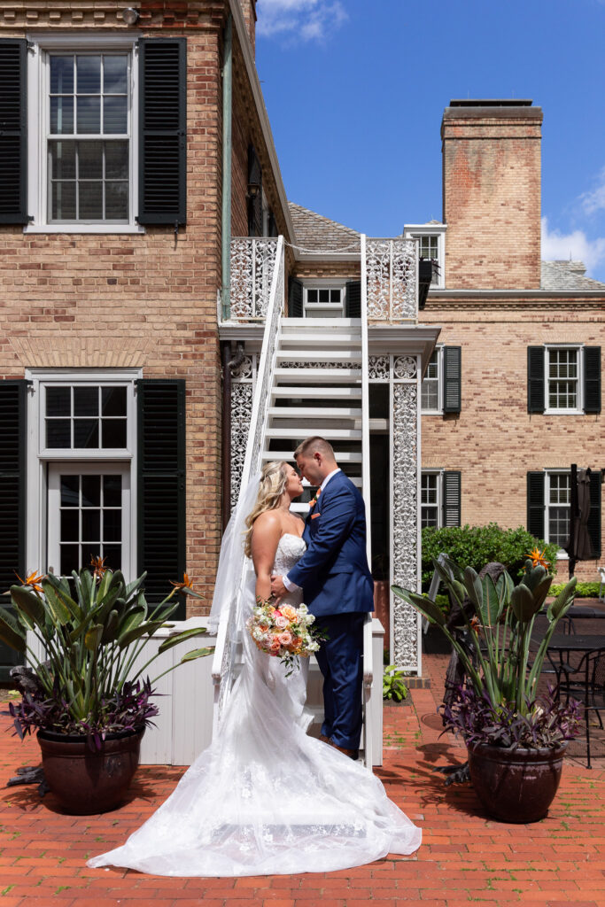The newlyweds embrace on a brick courtyard staircase, surrounded by elegant architecture and blooming planters during their Drumore Estates wedding portraits.