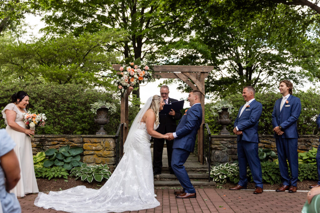 The couple laughs together during their wedding ceremony beneath a wooden arbor decorated with peach and blush flowers. Their wedding party smiles nearby as they hold hands in a joyful moment.