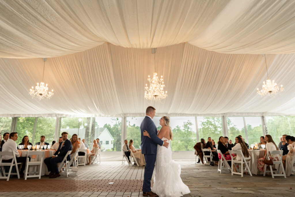 Bride and groom share their first dance in the center of the reception tent while wedding guests watch from their tables. Elegant drapery and chandeliers create a romantic atmosphere around the dance floor.