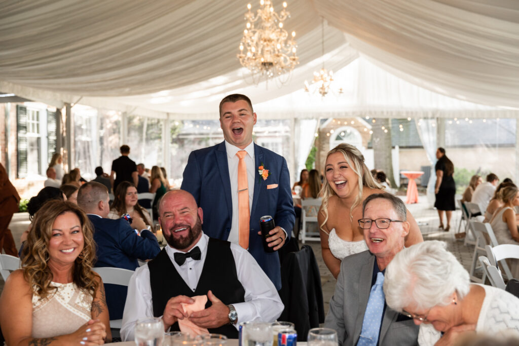 Newlyweds visit guests during the wedding reception, laughing with family members seated around a table beneath a draped tent ceiling and crystal chandeliers.