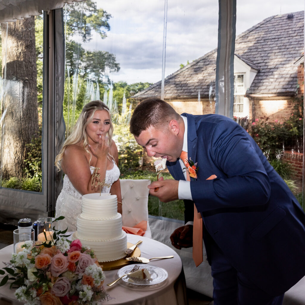 The groom takes an oversized bite of wedding cake while the bride licks the cake off of her fingers during their Lancaster Wedding cake cutting. Floral arrangements and candlelight decorate the sweetheart table beside them.