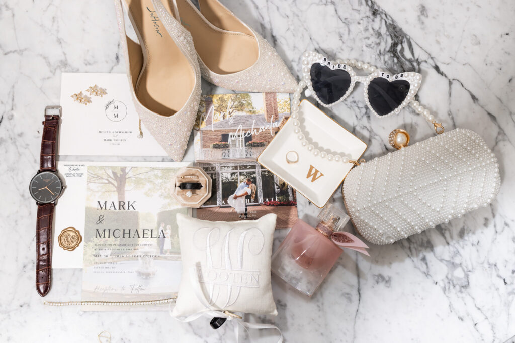 Wedding details including invitations, rings, shoes, perfume, a beaded clutch, and bridal accessories are carefully styled on a marble surface before the Drumore Estates wedding celebration.