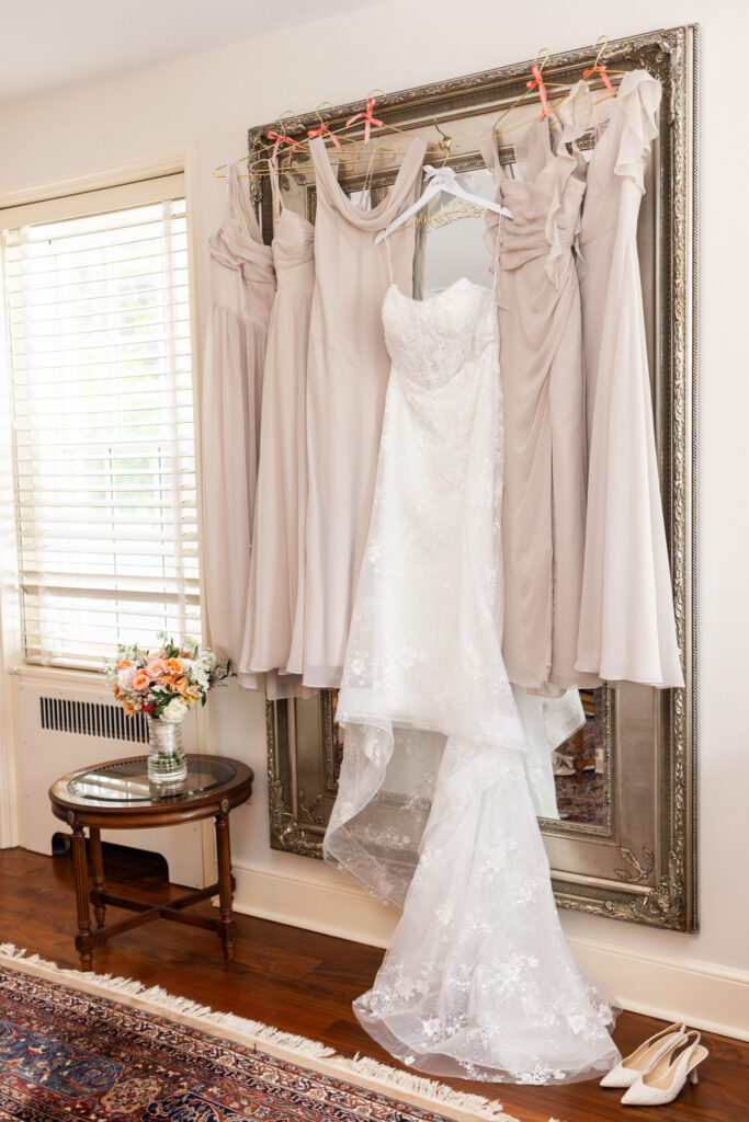 A lace wedding gown hangs beside blush bridesmaid dresses in front of an ornate mirror, with the bridal bouquet displayed nearby in a bright getting-ready suite.