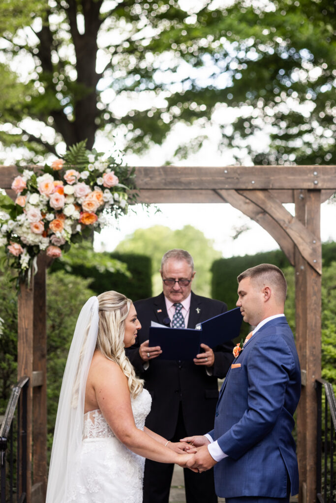 The bride and groom hold hands beneath a wooden arbor decorated with peach and ivory florals as they exchange vows during their outdoor Drumore Estates wedding ceremony.
