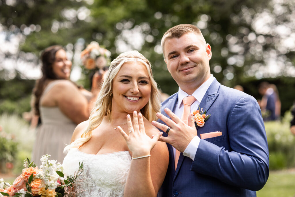 Newlyweds proudly display their wedding rings during a Lancaster Wedding portrait. The bride holds her bouquet while both smile toward the camera, showing off their rings against a softly blurred garden backdrop.