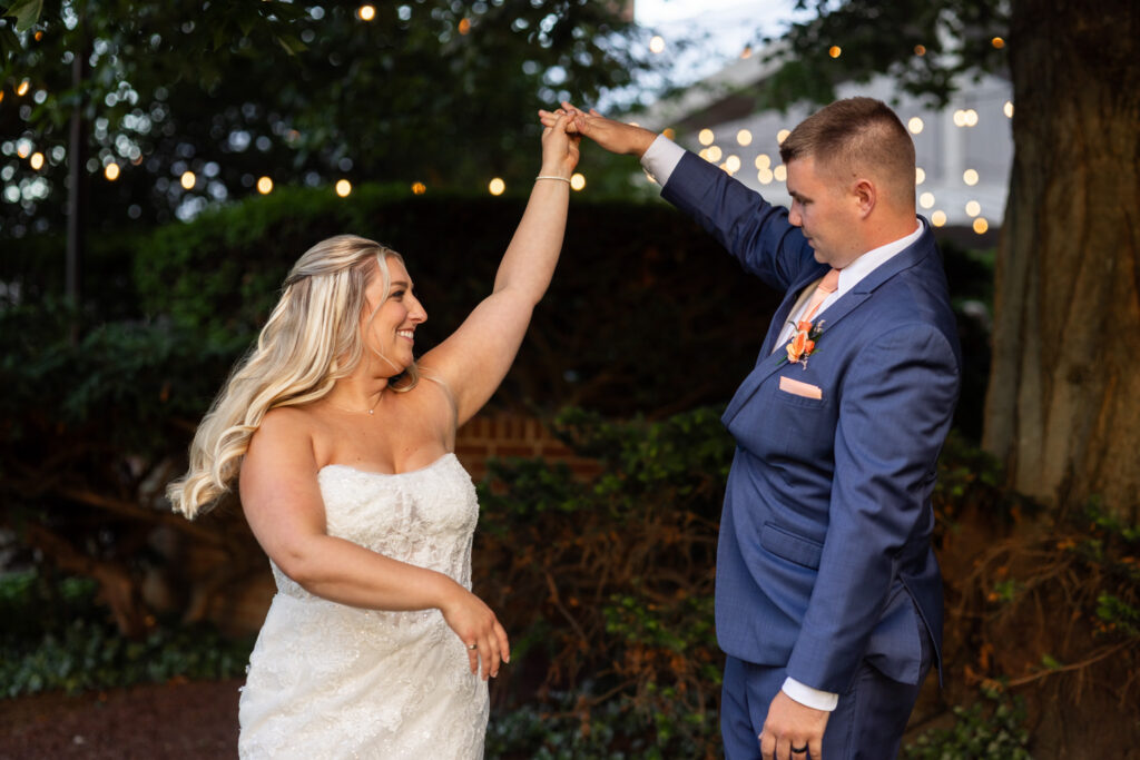 Bride twirls beneath her groom’s raised hand during a Lancaster Wedding portrait session at dusk. String lights glow softly in the background as the couple shares a playful moment outdoors.