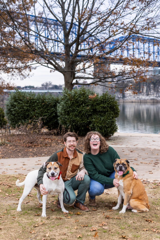 An engaged couple poses with their two dogs along a riverfront walkway with a blue bridge in the background. Both dogs wear festive bow ties while the couple kneels beside them, creating a joyful portrait that celebrates the entire family.