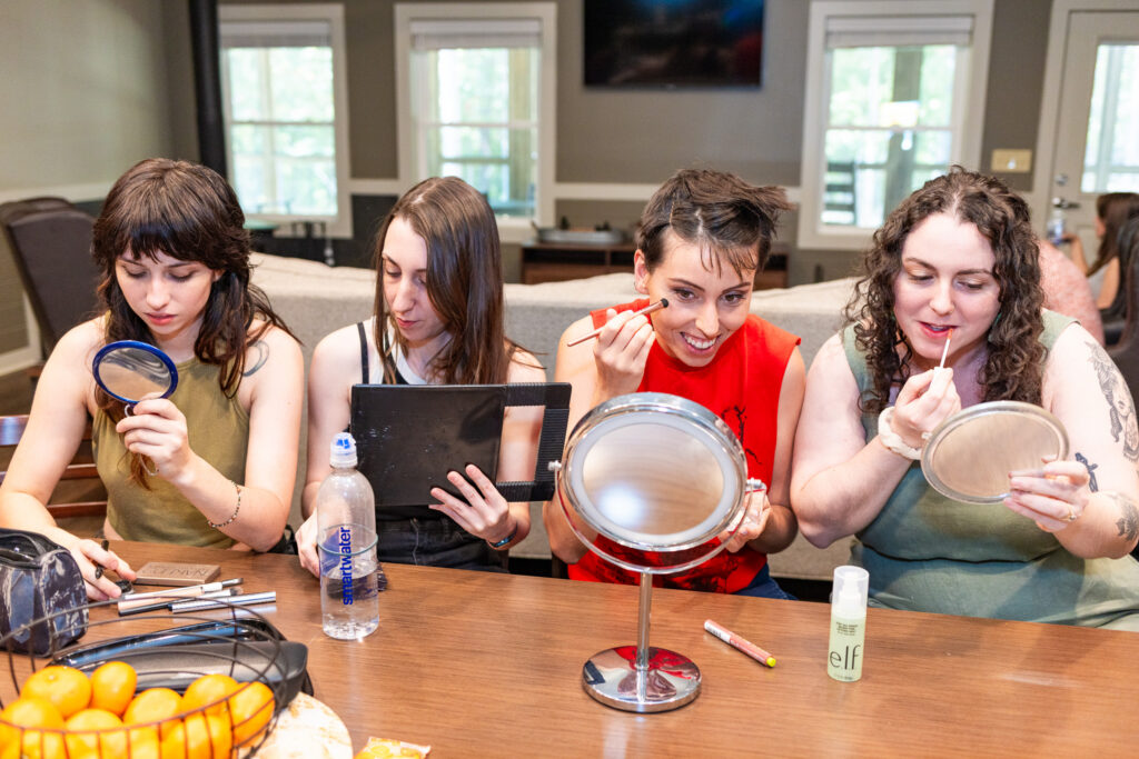 Four friends of various gender-fluidity sit around a table applying makeup and checking mirrors while getting ready together before a wedding ceremony. The best LGBTQ friendly wedding vendors create environments where wedding parties can show up exactly as they are