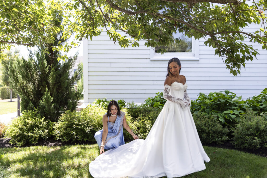 A bride in a white wedding gown stands on a lawn while her mom, in a light blue dress, arranges her train beside a white house.