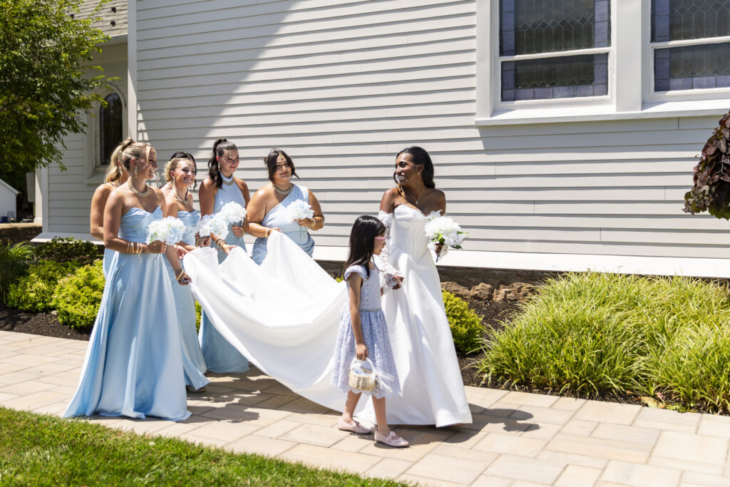 A bride in a white wedding dress walks beside a flower girl while four bridesmaids in light blue dresses hold up the bride’s train outside a white house.