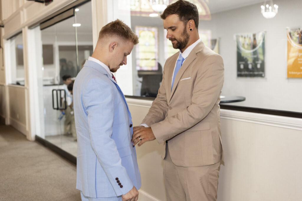 A groomsman in a tan suit helps the groom fasten his pale blue suit jacket before the New Jersey Wedding Chapel ceremony. The groom looks down as his friend adjusts the front of his jacket in a bright church hallway with glass doors and stained-glass windows visible in the background.