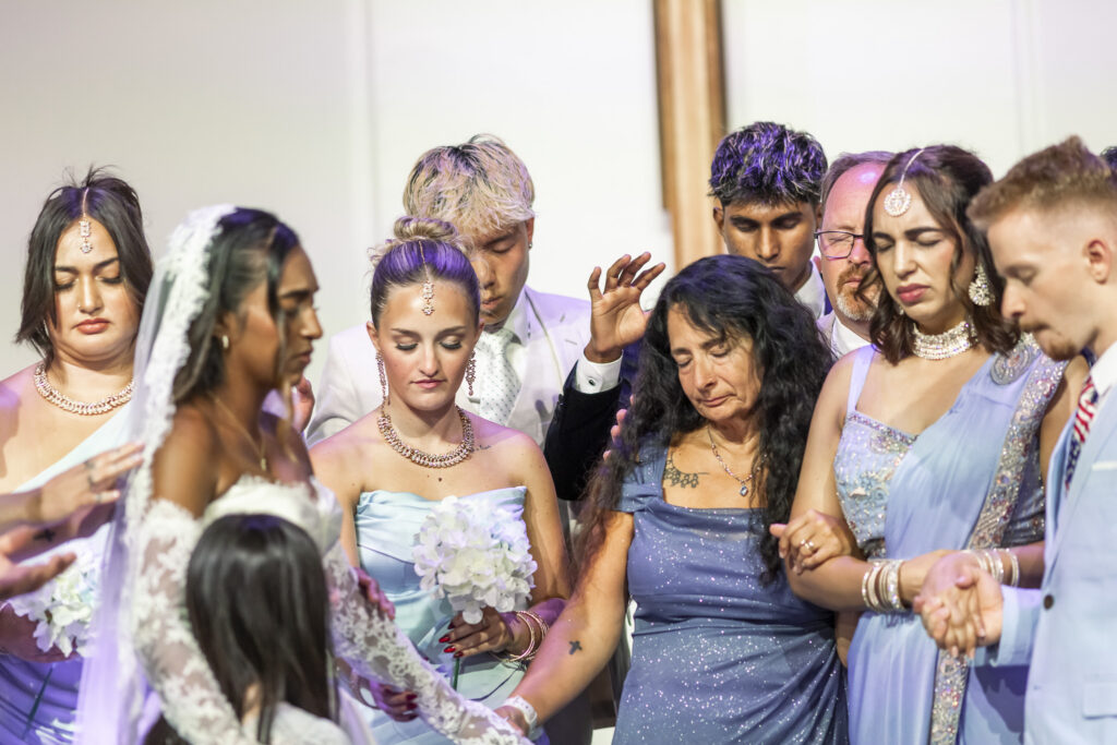 A bride and her family and wedding party gather closely around the altar in prayer during a New Jersey Wedding Chapel ceremony. With eyes closed and heads bowed, loved ones hold hands and reach toward the bride as they pray over the newly married couple.