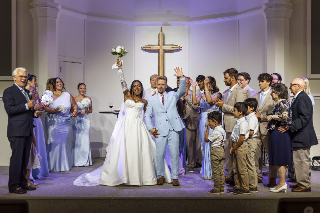 A newlywed couple stands hand in hand at the altar of a New Jersey Wedding Chapel, surrounded by cheering family and wedding party members. The bride raises her white bouquet overhead while the groom waves, with a large wooden cross centered on the church wall behind them.