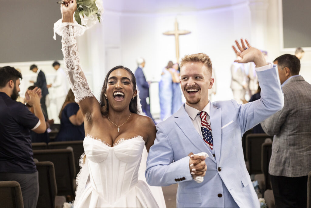 A joyful bride and groom walk back up the aisle after their New Jersey Wedding Chapel ceremony, holding hands and raising their free arms in celebration. The bride lifts her bouquet overhead while the groom smiles beside her in a pale blue suit and American flag tie.