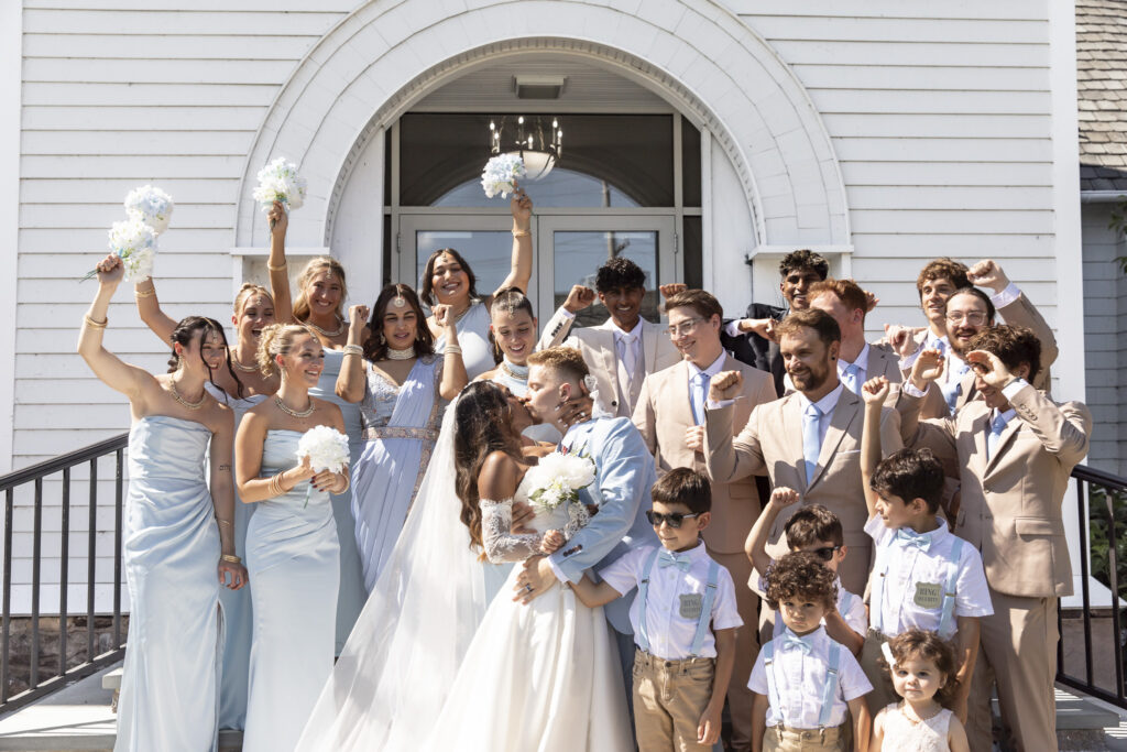 A bride and groom kiss outside a white New Jersey Wedding Chapel while their wedding party celebrates around them. Bridesmaids in pale blue dresses raise white bouquets and their arms in the air, while groomsmen in tan suits cheer behind them and several young ring bearers gather in front.