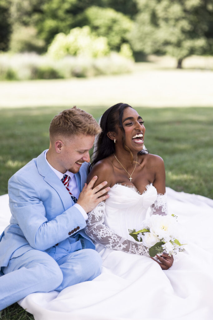 A bride and groom sit together on the grass laughing during portraits at their backyard wedding reception. The bride holds a white floral bouquet as her voluminous wedding gown spreads across the grass around the couple.