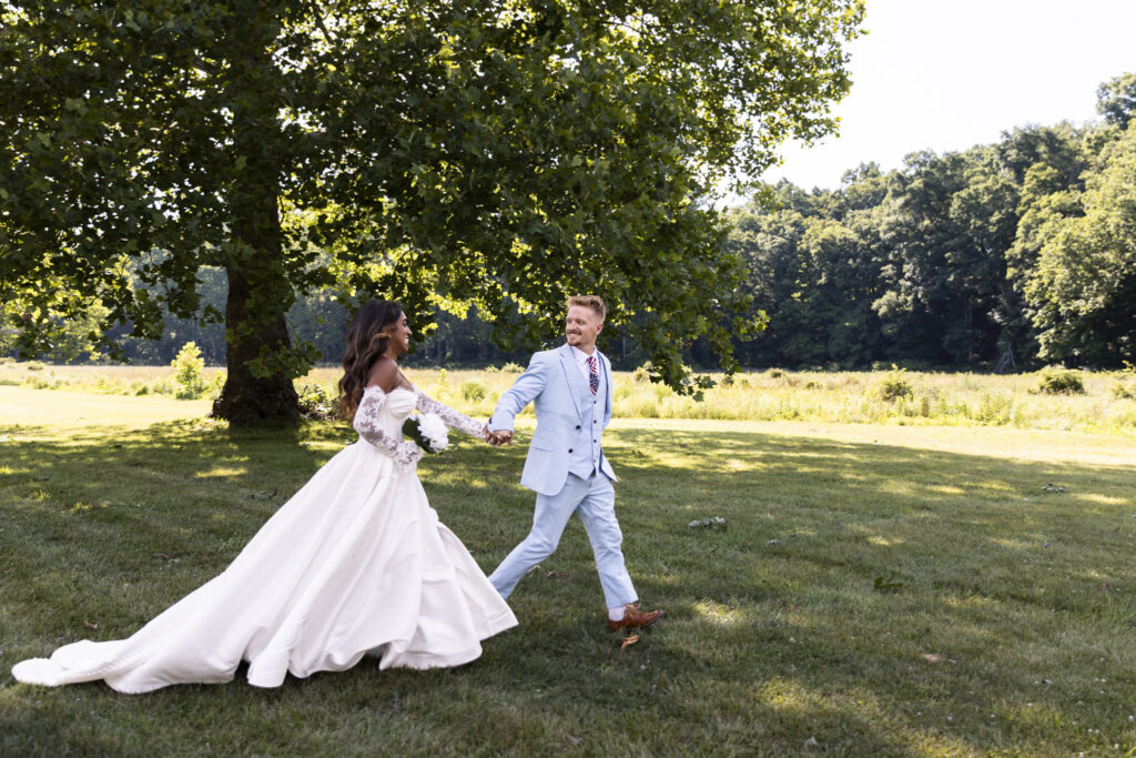 A newly married couple walks hand in hand across a sunlit lawn beneath a large leafy tree. The bride’s full white ball gown and long train sweep behind her as she looks toward the groom, who wears a pale blue three-piece suit and brown shoes. A bright meadow and dense green woodland stretch across the background.