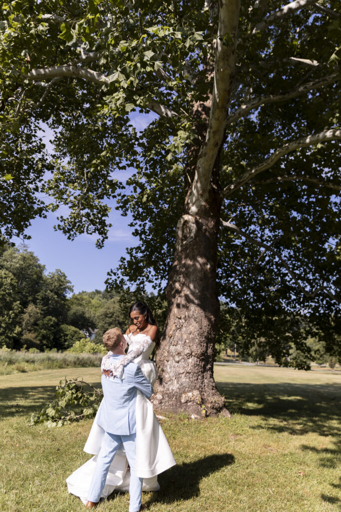 A bride and groom share a playful portrait beneath a towering shade tree, with the groom lifting his bride as they smile at one another. The expansive tree canopy and sunlit lawn create a natural, intimate setting around the newlyweds.