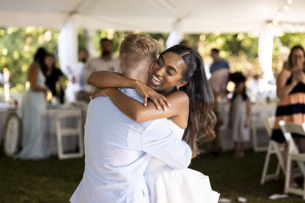 A bride and groom share a joyful embrace beneath a white reception tent, with the bride laughing as she wraps both arms around her new husband during their backyard wedding reception.