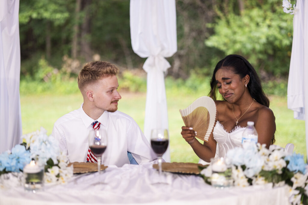 A groom looks affectionately toward his emotional bride as she holds a folding fan and wipes away tears during speeches at their backyard wedding reception. White and pale blue flowers, candles and wine glasses decorate the sweetheart table in front of them.