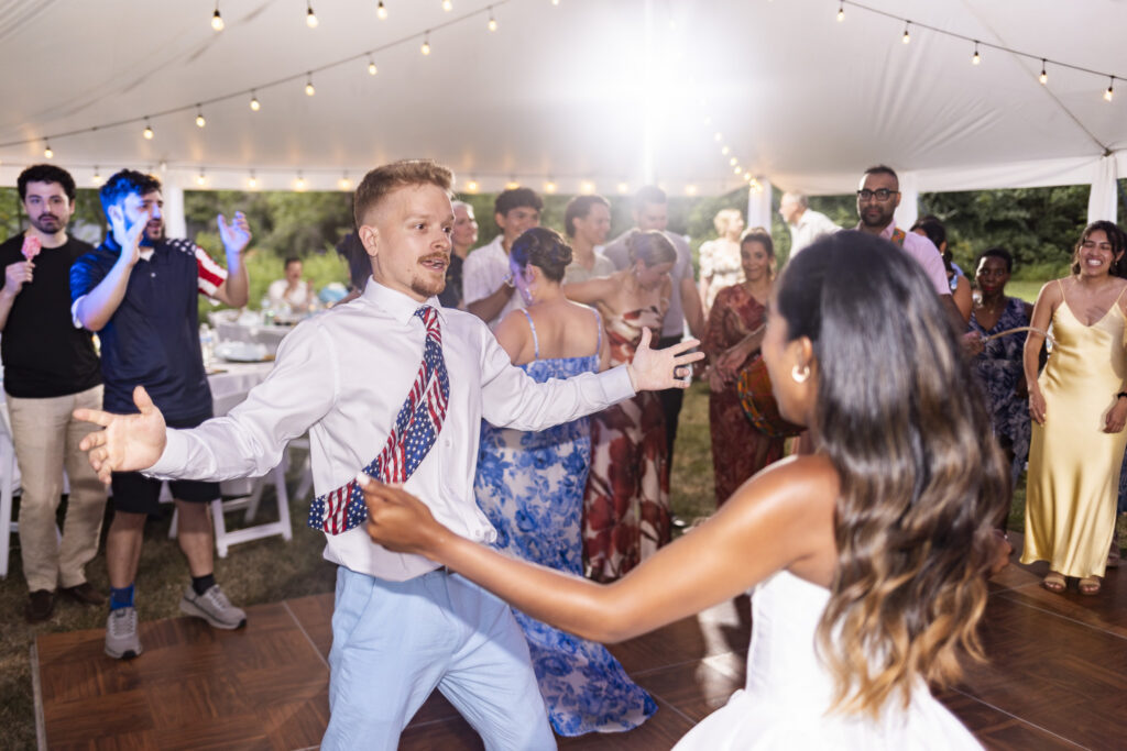 A groom in light blue trousers and a white dress shirt reaches his arms toward the bride while dancing at their backyard wedding reception. The bride is seen from behind with long dark hair as the couple faces one another on the wooden dance floor. Guests dance and watch beneath the softly lit white reception tent.