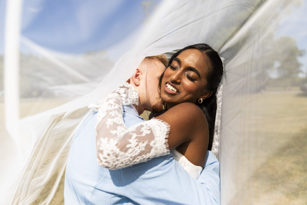 A close-up wedding portrait captures the bride smiling with her eyes closed as she embraces the groom beneath her flowing veil. Her lace-covered arms wrap around his shoulders while layers of translucent white fabric billow around the couple, framing them against a softly blurred outdoor landscape. An intimate image following their New Jersey wedding chapel nuptuals