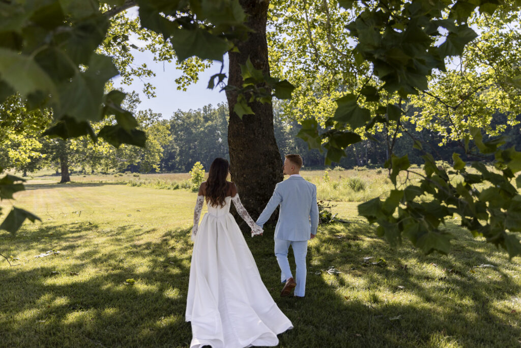 A newlywed couple walks hand in hand beneath a large shade tree during their backyard wedding reception, surrounded by a sunlit lawn and lush green landscape. The bride’s full white gown trails behind her as the groom walks beside her in a pale blue suit.