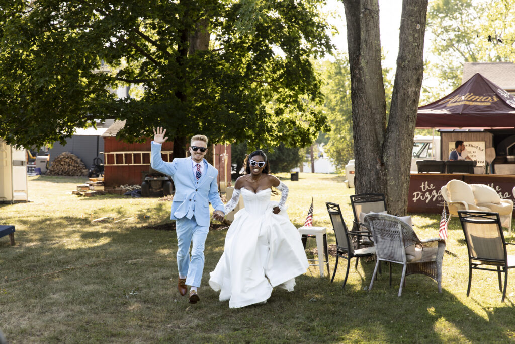 A smiling bride and groom walk hand in hand across the lawn during their backyard wedding reception, with the groom waving toward the camera and the bride wearing playful white heart-shaped sunglasses. Large shade trees, casual seating and an outdoor pizza station fill the sunny backyard behind them.