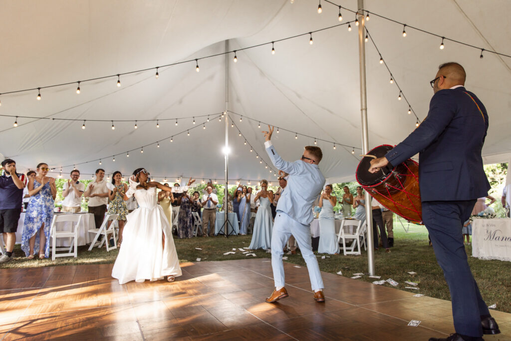 A bride and groom dance several feet apart on a wooden dance floor as a live Indian drummer plays beside them. The bride reaches toward the groom while he raises one hand in the air, and guests gather around the dance floor clapping and cheering beneath a white tent filled with strands of warm lights.